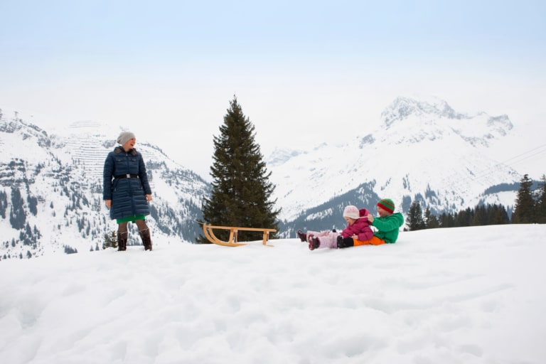 Eine Frau zieht einen Schlitten, zwei Kinder sitzen neben dem Schlitten und lachen, dahinter verschneite Bergkulisse Arlberg