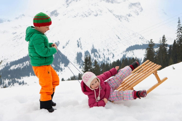 Bub zieht Mädchen mit dem Schlitten, Mädchen ist halb vom Schlitten gefallen, beide lachen; dahinter verschneite Arlberg-Kulisse
