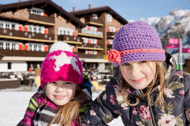 Zwei Mädchen mit bunter Winterbekleidung sitzen vor dem Familienhotel Sonnenburg in der Sonne, sie lächeln in die Kamera
