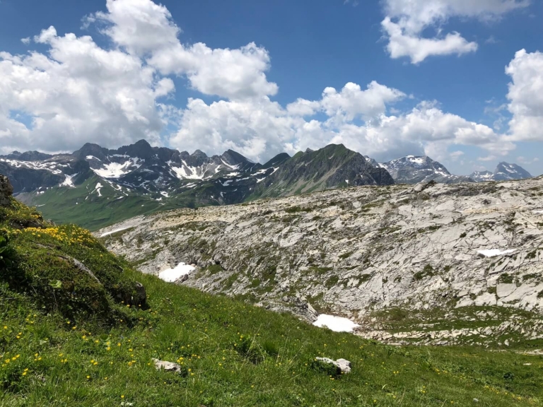 Blick auf das Gestein beim Geoweg Rüfikopf: Grüne Wiese im Vordergrund, raues Gestein und Berge dahinter