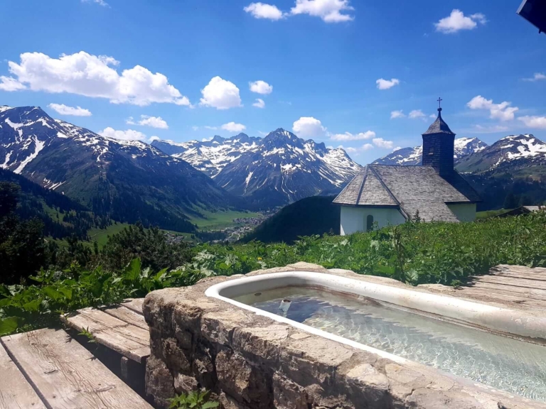 Blick auf die Kapelle von Bürstegg; im Vordergrund ist ein steinerner Brunnen zu sehen, im Hintergrund ragen beschneite Berggipfel in den strahlend blauen Himmel.