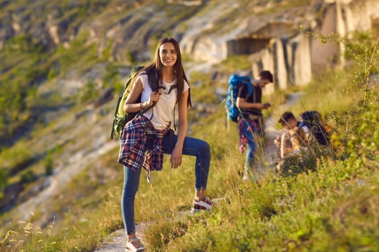 Drei Teenager beim Wandern in den Bergen; zwei Jugendliche befinden sich im Hintergrund, eine Teenagerin blickt direkt in die Kamera und lächelt