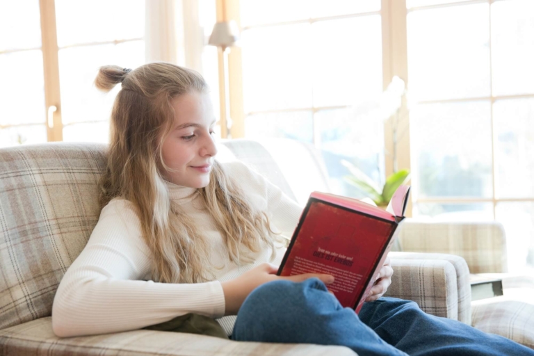 Eine Teenagerin mit weißem Rollkragenpulli und langen blonden Haaren sitzt in einem gemütlichen Sessel und liest ein rotes Buch in der Sonnenburg