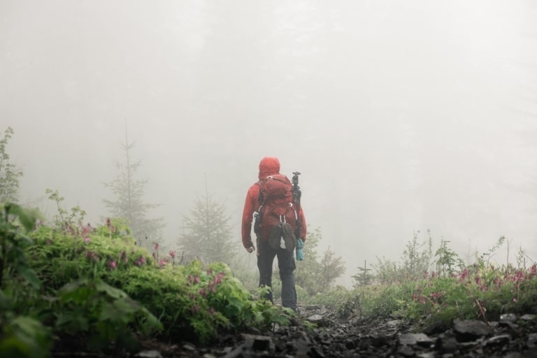 Mann mit roter Regenjacke und rotem Rucksack wandert durch eine nebelige Almlandschaft