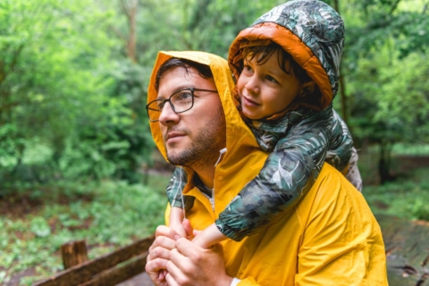 Aktivitäten_Sommer_Wandern_Regen_Vater Kind Vater und Sohn tragen eine Regenjacke mit Kapuze; der Sohn hängt sich mit seinen Armen über die Schultern des Vaters. Beide sind nass. Im Hintergrund sind saftig grüne Bäume zu sehen.