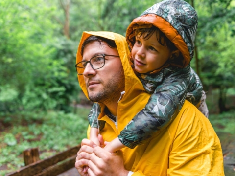 Vater und Sohn tragen eine Regenjacke mit Kapuze; der Sohn hängt sich mit seinen Armen über die Schultern des Vaters. Beide sind nass. Im Hintergrund sind saftig grüne Bäume zu sehen.