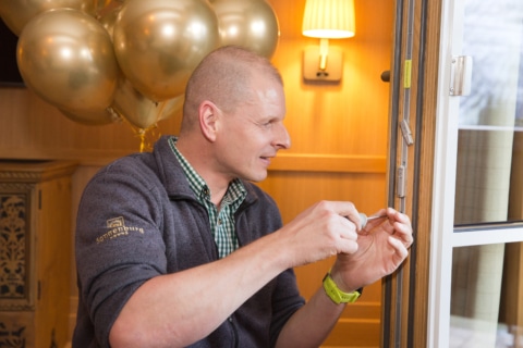 Sonnenburg_Familie_Mitarbeiter_Haustechnik_Tür_einstellen_Zimmer_2022_Ballons (1) Ein Handwerker bei der Wartung einer Fensterverriegelung in einem Hotelzimmer