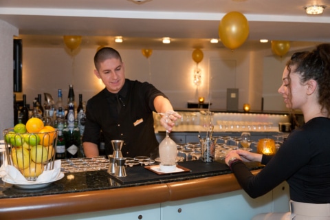 Sonnenburg_Familie_Mitarbeiter_Bar_Almin_Cocktail_mixen_Sommer_2024 (16) Barkeeper mixt einen Cocktail für eine Kundin an der eleganten Bar