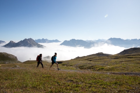 Sonnenburg_Aktivitaeten_Sommer_Wandern_RichtungMohnenfluh_Frauen_InDerFerne_Nebel_Sonne_Wiese_Berge (7) Zwei Wanderer auf dem Weg zum Gipfel