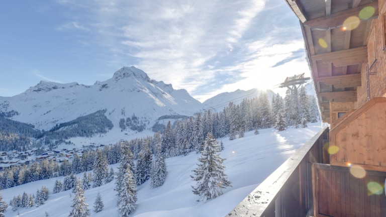 Panoramaansicht der schneebedeckten Alpen mit dichten Föhrenwäldern und traditionellen Chalets im Tal.