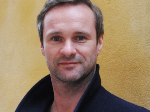 ClemensBerger Ein professioneller Mann in eleganter Oberbekleidung, steht vor einer gelben Wand, symbolisiert die Aufgeschlossenheit und Gastfreundschaft in der Tourismus- und Gastronomiebranche.