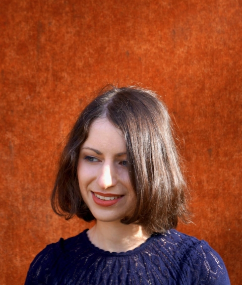 Sarah-Kuratle-(c)privat Junge Frau mit braunen Haaren, die entspannt vor einer orangefarbenen Wand steht, ideal für die Darstellung des Tourismus und der Gastronomie.